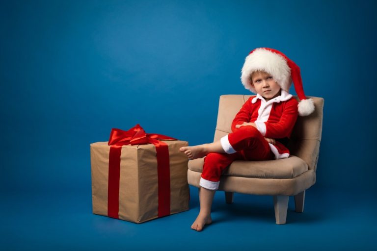 Angry kid sitting and wearing Santa costume with a Christmas gift on his side