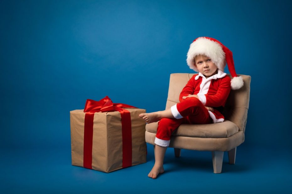 Angry kid sitting and wearing Santa costume with a Christmas gift on his side