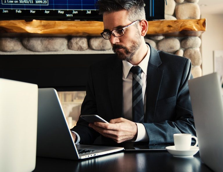 man in suit in front of a laptop searching for executive resumes online while holding a smartphone